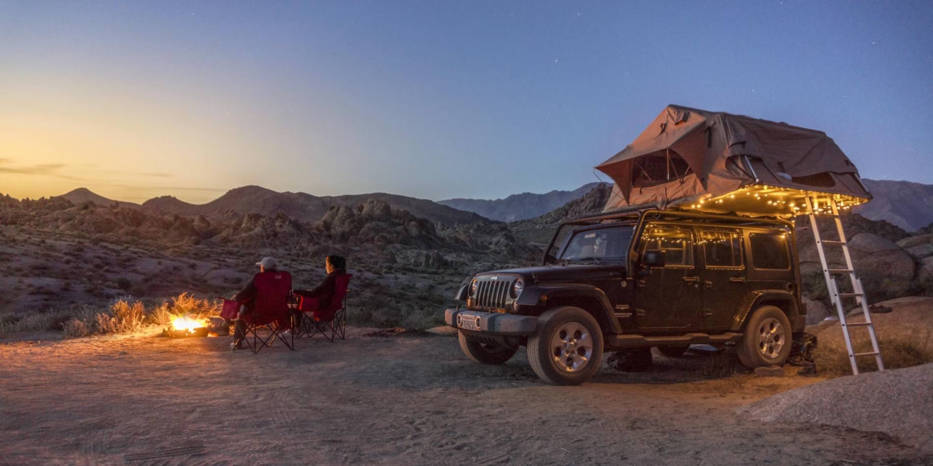 Overlanding and 2 men sitting near a campfire and an SUV with a tent atop parking behind the two men